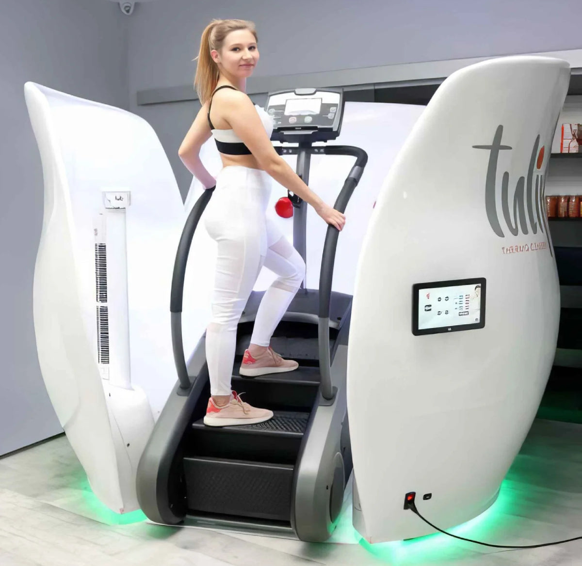 A woman in a black and white sports bra and white leggings is stepping on a treadmill-style exercise machine inside a white room. The machine has curved white side panels with a digital display screen.