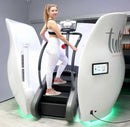 A woman in a black and white sports bra and white leggings is stepping on a treadmill-style exercise machine inside a white room. The machine has curved white side panels with a digital display screen.