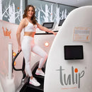 A woman in a white sports bra and leggings stands on a white Tulip Thermo Climber treadmill machine in a fitness center. The machine has digital screens and the brand name 'tulip' printed on the side.