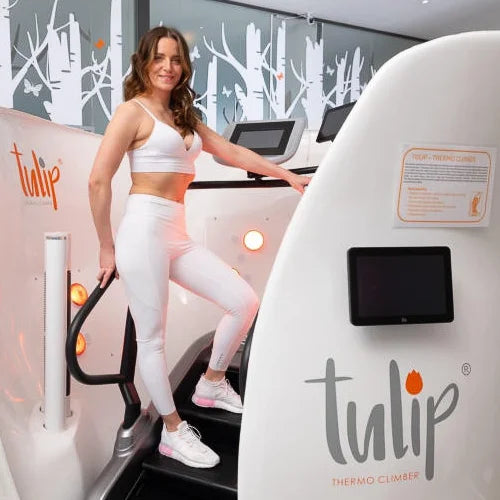 A woman in a white sports bra and leggings stands on a white Tulip Thermo Climber treadmill machine in a fitness center. The machine has digital screens and the brand name 'tulip' printed on the side.