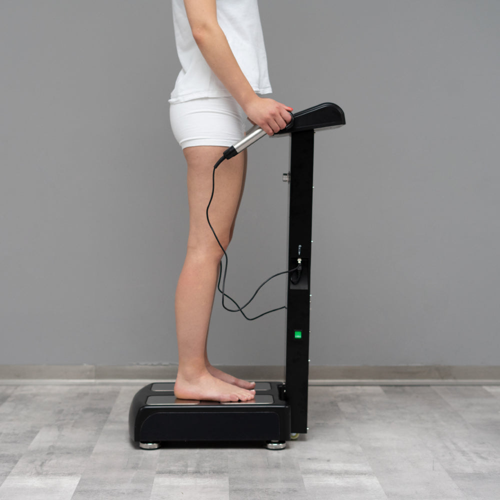 A person standing barefoot on a black body composition machine, holding the handle with one hand, in a room with a gray wall and light gray floor.