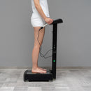 A person standing barefoot on a black body composition machine, holding the handle with one hand, in a room with a gray wall and light gray floor.