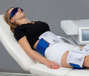 A woman reclines on a white treatment table wearing a blue eye mask and a white compression garment with blue support pads attached to white electronic devices. A medical-style machine is visible beside her against a gray studio background.