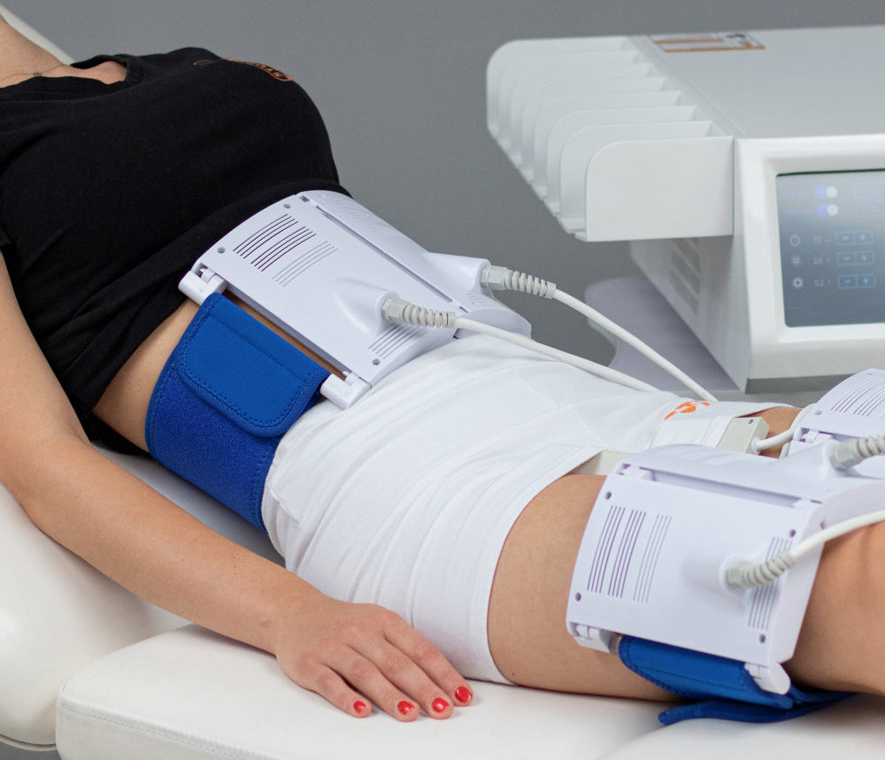 A person reclining on a treatment table with a waist and thigh massage or electrical stimulation device in use. The white and blue device is strapped around the waist and upper thigh, with cables connected to a control unit on a gray background.