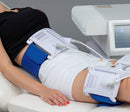 A person reclining on a treatment table with a waist and thigh massage or electrical stimulation device in use. The white and blue device is strapped around the waist and upper thigh, with cables connected to a control unit on a gray background.