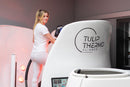 A woman in a white athletic outfit rides a white Tulip Thermo Climber exercise machine in a modern indoor facility. The machine features the black printed logo 'TULIP THERMO CLIMBER' on its curved side panel.