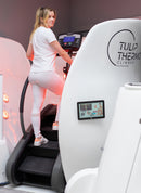 A woman in a white athletic outfit uses a treadmill inside a white and black medical-style chamber with visible red light therapy panels. The machine has the brand name 'TULIP THERMO CLIMBER' printed on the side.